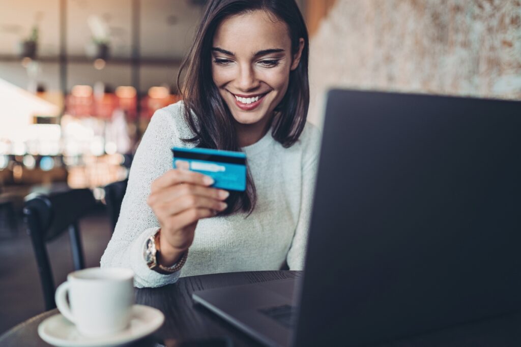 Uśmiechnięta kobieta z kartą kredytową siedzi przed laptopem. Zakupoholizm - objawy